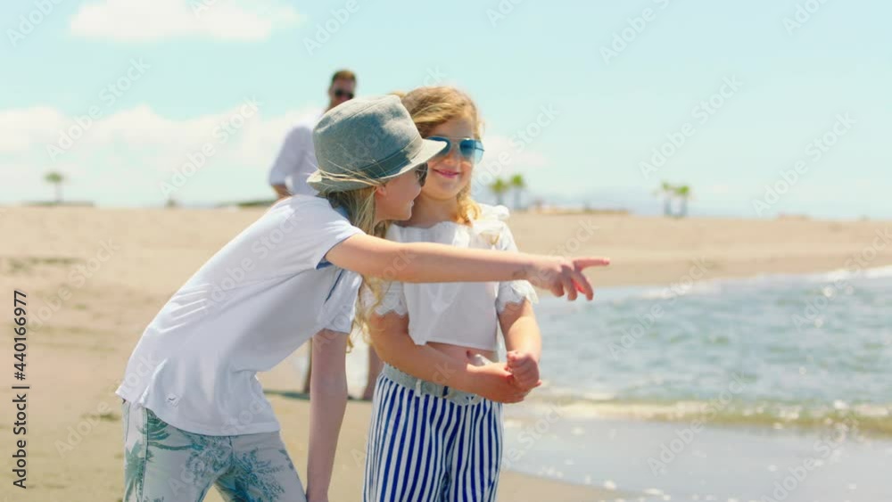 Children in summer clothing wearing sunglasses standing on beach. Boy explaining something to girl and pointing by hand forward. Sunny summer day at seaside.