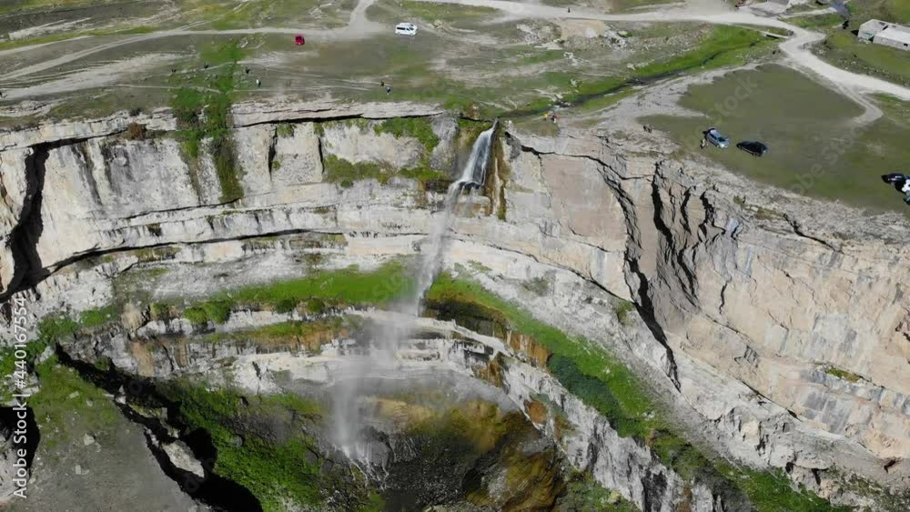 Aerial view of a high waterfall in Dagestan. Water rushes down the cliff in sunlight. The stone plateau is covered with greenery and roads where cars drive. Rainbow reflections from water Aerial view of a high waterfall in Dagestan. Water rushes down the cliff in sunlight. The stone plateau is covered with greenery and roads where cars drive. Rainbow reflections from water