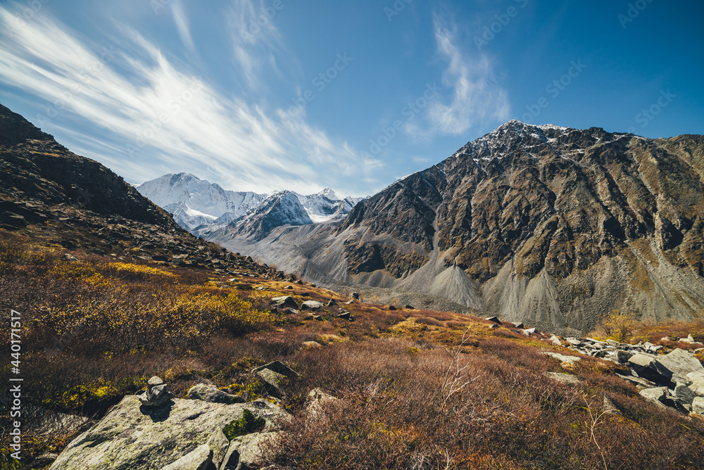 Autumn sunny landscape with highland valley and snow-covered high mountains under blue cloudy sky with cirrus clouds. Atmospheric mountain scenery with sunlit valley among rocky mountains in autumn.