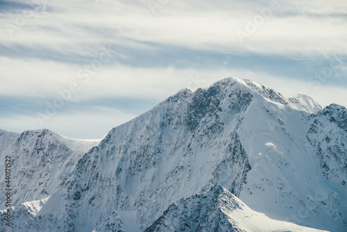 Great view to high snowy mountain wall with peaked top under cirrus clouds in sky. Alpine landscape with big snow covered mountains with sharp pinnacle in sunshine. White-snow pointy peak in sunlight.