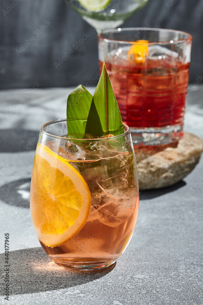 Orange Gin tonic cocktail for lifestyle design. Summer cocktail concept background. Gin tonic drink with orange slice and banana leaf. Cocktail glass on vintage table with sunshine and harsh shadow. Orange Gin tonic cocktail for lifestyle design. Summer cocktail concept background. Gin tonic drink with orange slice and banana leaf. Cocktail glass on vintage table with sunshine and harsh shadow.