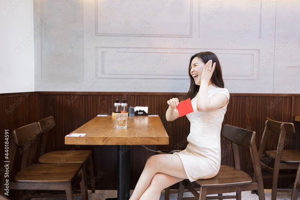Beautiful young asian woman in traditional white dress named cheongsam sitting in Japanese restaurant or cafeteria and waiting ordered food. Girl holding red envelope. Restaurant interior decorated Beautiful young asian woman in traditional white dress named cheongsam sitting in Japanese restaurant or cafeteria and waiting ordered food. Girl holding red envelope. Restaurant interior decorated