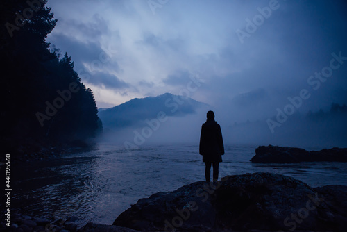 Human silhouette in a blue haze on the background of mountains and river. Thick fog in the evening twilight. Mysterious atmosphere. Concept of solitude, reflection, meditation.