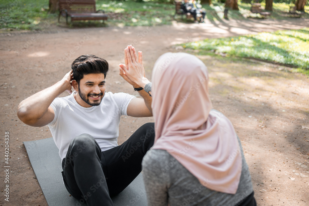 Obraz premium Muscular hindu guy training abdominal muscles on yoga mat with help of woman in hijab that holding his feet. Active young couple training together on fresh air.
