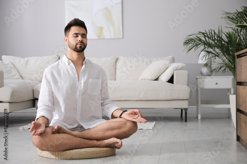 Young man meditating on straw cushion at home, space for text