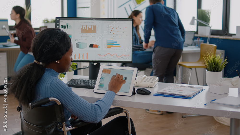 Obraz premium Black handicapped woman entrepreneur with locomotor disabilities using computer and tablet in same time working in business start-up office sitting in wheelchair. Busy multitasking african employee