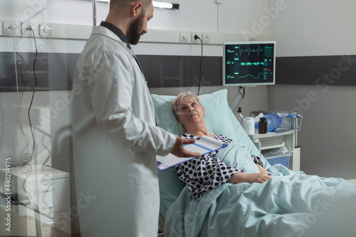 Sick senior woman lying in hospital bed listening doctor talking about treatment and diagnosis. Patient inhale and exhale with help from oxygen tube. Iv drip bag attached on arm. photo