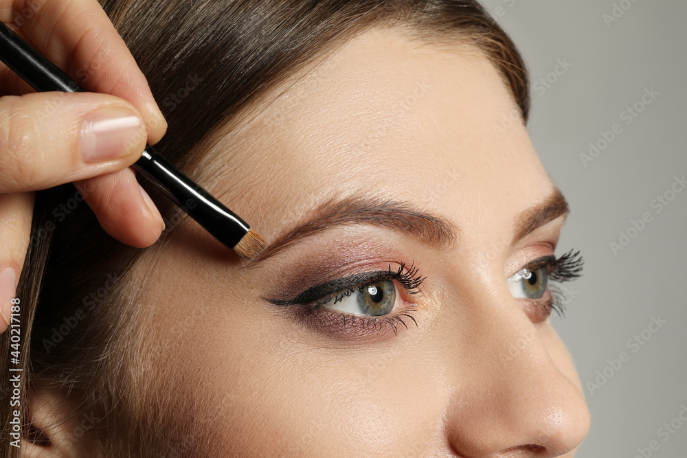 Artist correcting woman's eyebrow shape with powder on grey background, closeup