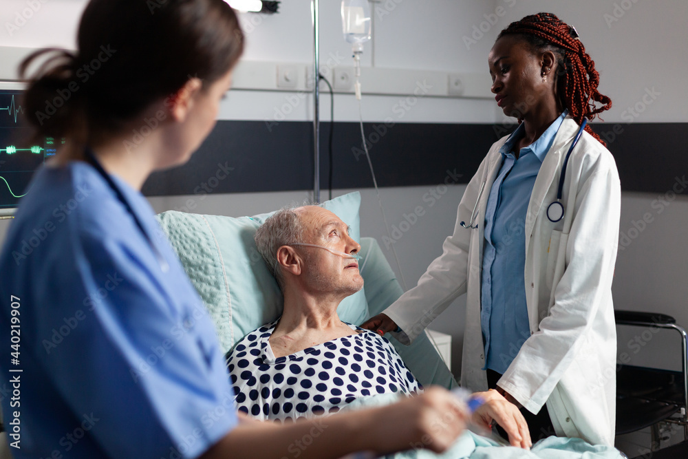Obraz premium African doctor ward in hospital room discussing with sick senior man laying in bed breathing through oxygen tube with iv dirp bag attached, during consultation. Medical nurse taking notes on clipboard