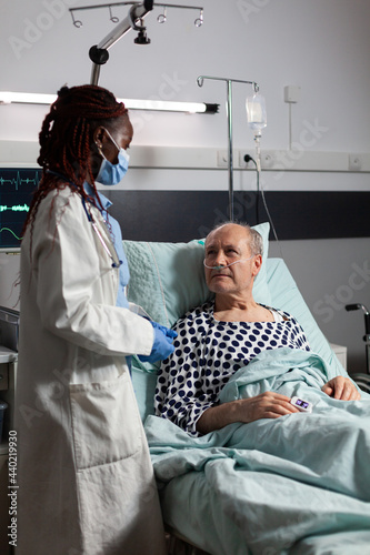 African american medical practitoner wearing chirurgical mask, in hospital room during consultation of sick unwell senior man, breathing with help from test tube and iv drip attached. photo