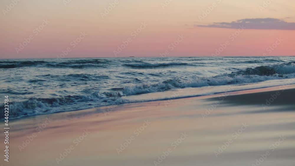 Low angle view of sea waves rolling and splashing on sandy beach. Pink twilight sky. Summer evening at seaside.