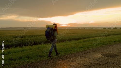 Active healthy caucasian woman with backpack walks along road in rays of sunset. Young woman traveler hiking in summer nature. The traveler travels in the countryside, meditation, ecotourism, hiking.