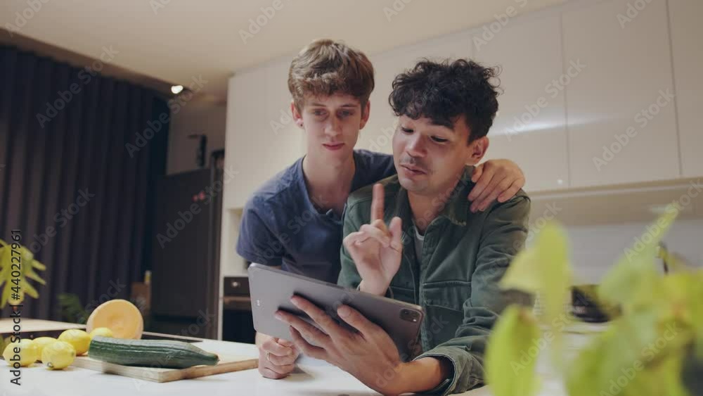 Romantic male couple bonding in kitchen making healthy meal searching recipe on digital tablet