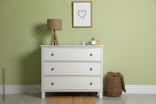 Modern white chest of drawers with lamp and decor near light green wall indoors © New Africa