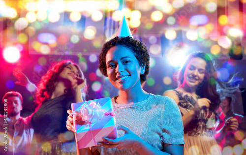 birthday, celebration and nightlife concept - happy smiling young african american woman in party cap with gift box in ultraviolet neon lights over nightclub background