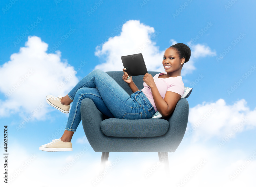 cloud computing, technology and people concept - happy smiling young african american woman with tablet pc computer sitting in modern armchair over blue sky background