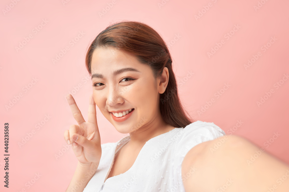 Young cheerful woman taking a self-portrait and gesturing v-sign against pink background Young cheerful woman taking a self-portrait and gesturing v-sign against pink background