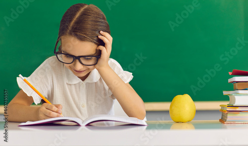 Foto Concentrated schoolgirl child writing in copy-book while sitting at desk with apple and stationary study supplies against green chalkboard