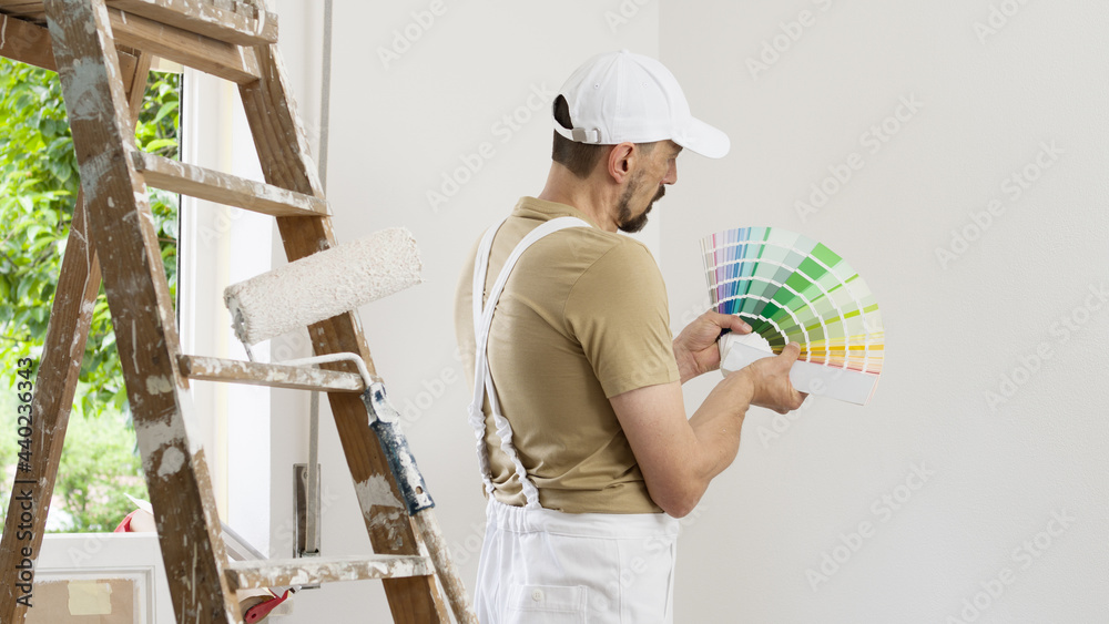 house painter man decorator choose the color using the sample swatch, work the white wall of the house to renovate, a wooden ladder, a paint roller and a green window as a background