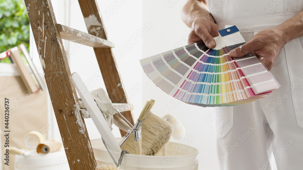 hands of house painter man decorator choose the color using the sample swatch, work of the house to renovate, a wooden ladder with paint brushes and a bucket as a background, close up - obrazy, fototapety, plakaty 