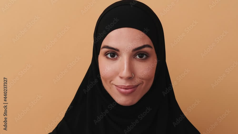 A close-up view of a beautiful arabian woman wearing a black hijab is looking to the camera standing isolated over a beige wall in the studio A close-up view of a beautiful arabian woman wearing a black hijab is looking to the camera standing isolated over a beige wall in the studio