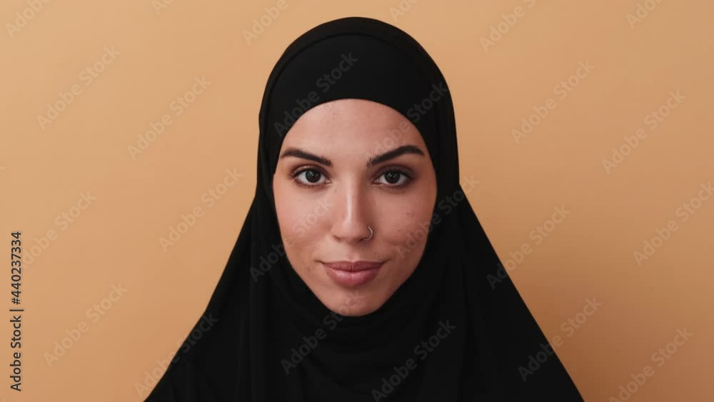 A close-up view of a beautiful arabian woman wearing a black hijab is posing to the camera standing isolated over a beige wall in the studio A close-up view of a beautiful arabian woman wearing a black hijab is posing to the camera standing isolated over a beige wall in the studio