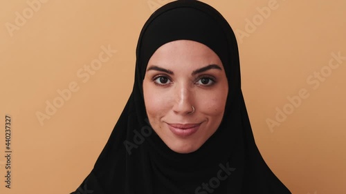 A close-up view of a beautiful arabian woman wearing a black hijab is looking to the camera standing isolated over a beige wall in the studio photo