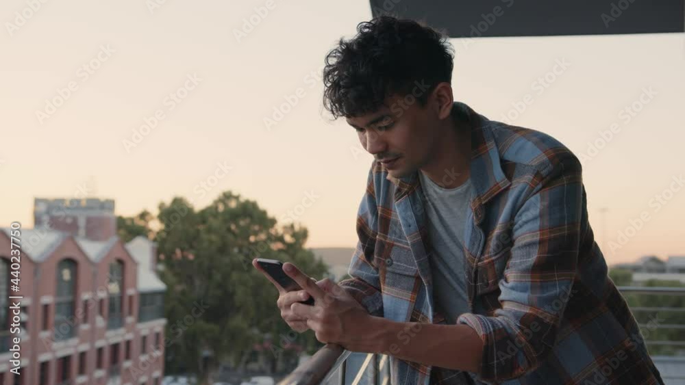 Mixed race male texting on cellular device messaging friends while leaning over balcony