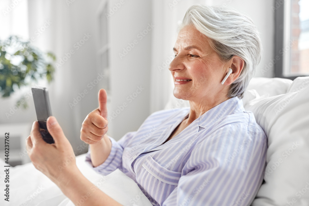 technology, old age and people concept - senior woman with smartphone and wireless earphones having video call in bed at home and showing thumbs up