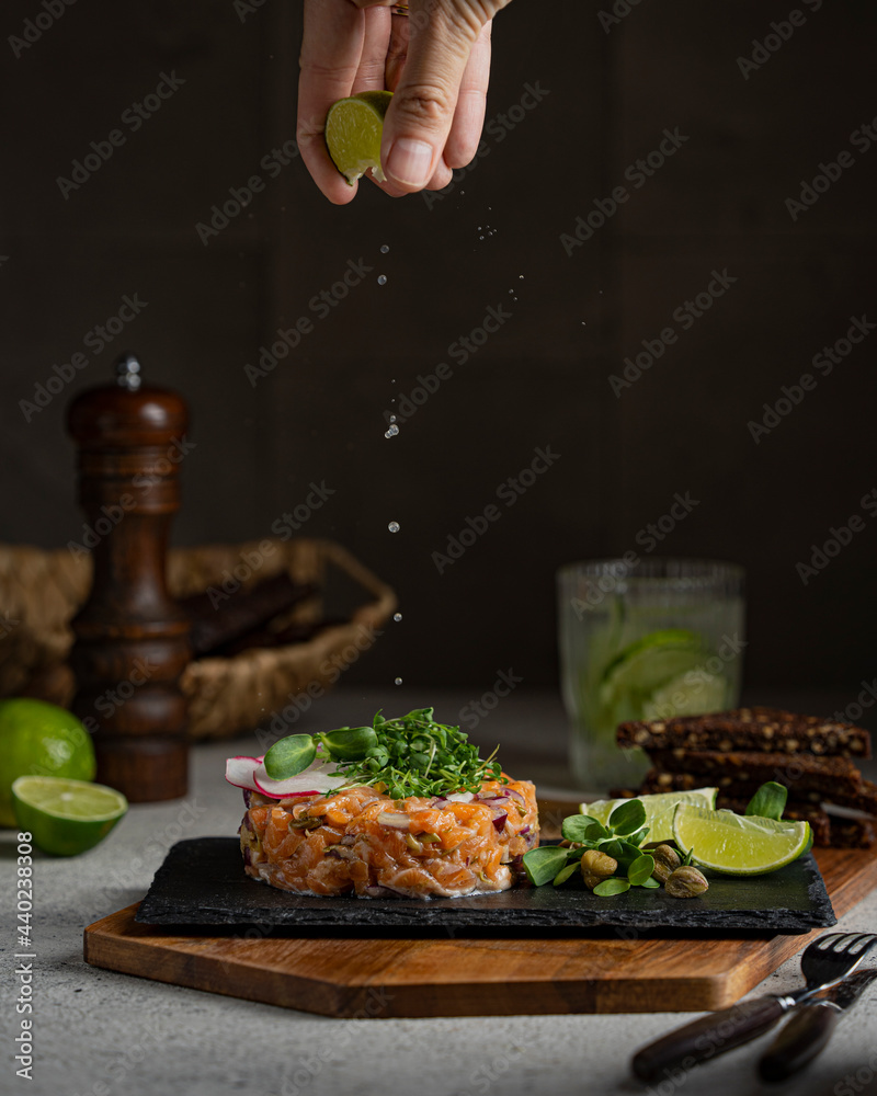 Salmon fish tartare with micro greens, lime on black stone plate, dark background. Grey textured table, healthy sea food concept. Macro shot with copy space.