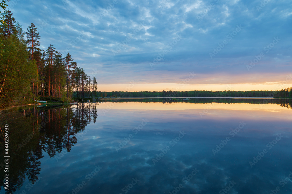 Beautiful view of quiet forested shoreline, cloudy sky and their reflections on a calm lake at sunset in Finland at summer.