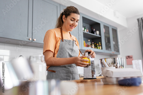 culinary, baking and people concept - happy smiling young woman cooking food on kitchen at home and grating lemon peel on grater photo