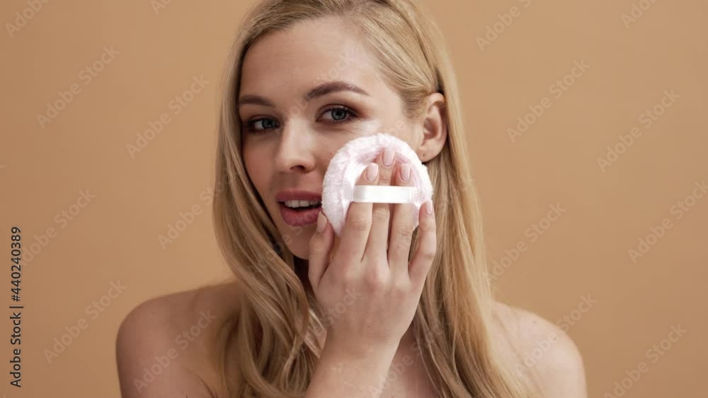A pleased blonde woman is applying powder brush on her face using powder sponge while standing isolated over a beige wall in the studio A pleased blonde woman is applying powder brush on her face using powder sponge while standing isolated over a beige wall in the studio