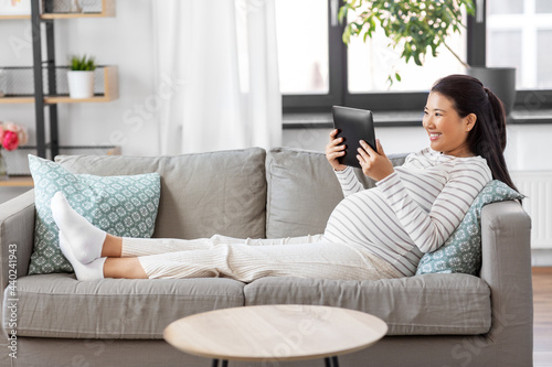 pregnancy, rest, people and expectation concept - happy smiling pregnant asian woman with tablet pc computer sitting on sofa at home