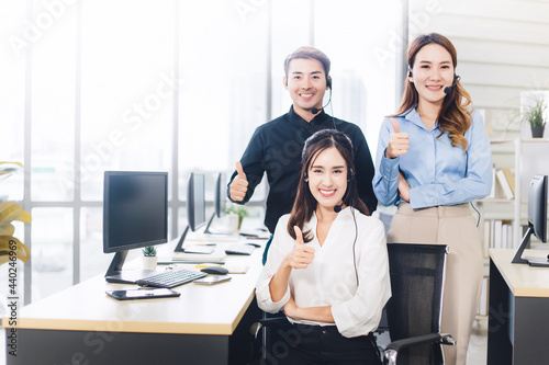 Asian customer service team looking at the camera and doing thumb up in call center. Group of young smiling operator with headset in modern office. Copy space