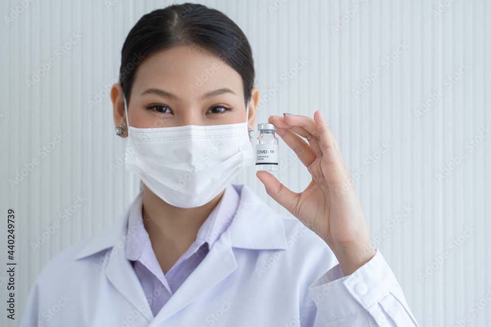 Selective focus on Covid-19 vaccines vial bottle in hand of Asian young doctor woman wear face mask, white gown and smiling on white background. Close up portrait Selective focus on Covid-19 vaccines vial bottle in hand of Asian young doctor woman wear face mask, white gown and smiling on white background. Close up portrait