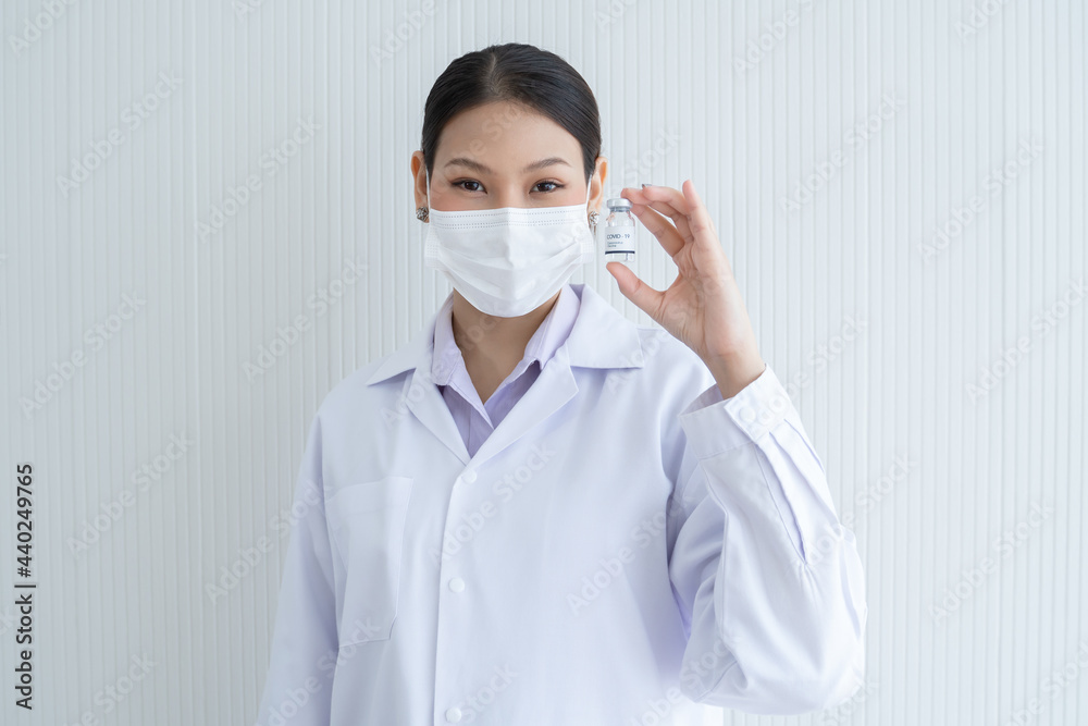 Portrait of Asian young doctor or scientist woman holding showing Covid-19 vaccines vial bottle in hand wear face mask, white gown and smiling. White background Portrait of Asian young doctor or scientist woman holding showing Covid-19 vaccines vial bottle in hand wear face mask, white gown and smiling. White background