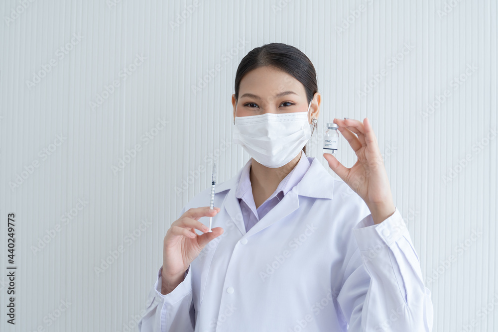 Portrait of Asian young doctor or scientist woman holding showing Covid-19 vaccines vial bottle in hand with face mask, white gown and holding syringe in other hand. White background. Portrait of Asian young doctor or scientist woman holding showing Covid-19 vaccines vial bottle in hand with face mask, white gown and holding syringe in other hand. White background.