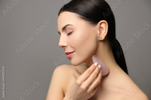 Beautiful young woman doing facial massage with gua sha tool on grey background  closeup