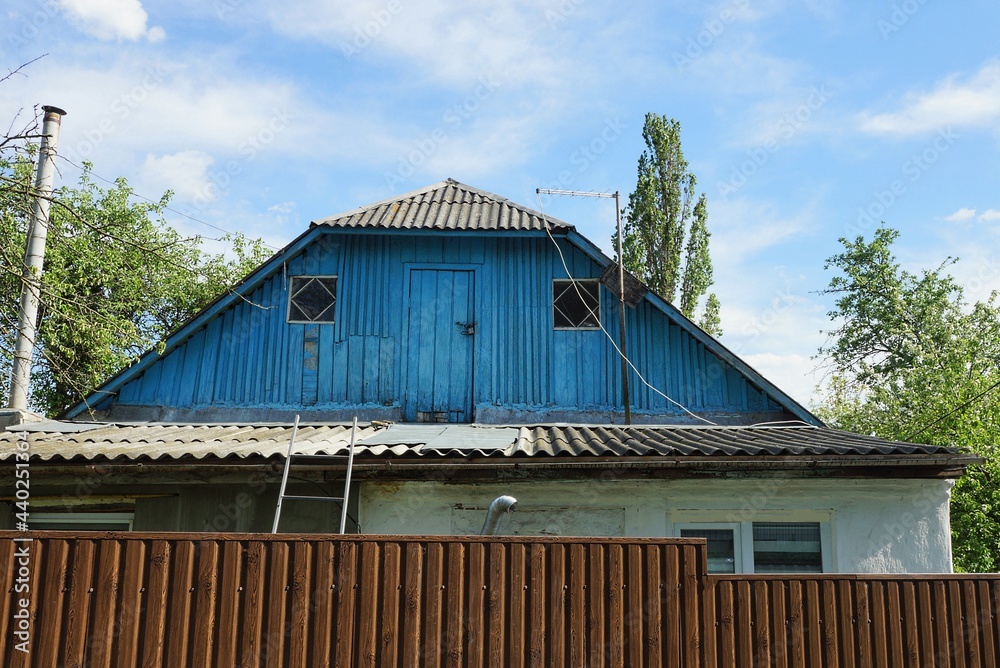 blue old wooden loft of a rural house with a door and small windows behind a brown fence against the sky