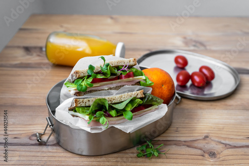 Sandwich mit Salatblätter und Schinken in einer wiederverwendbaren Blechdose, Orangensaft und Cocktailtomaten auf einem Holz Tisch.