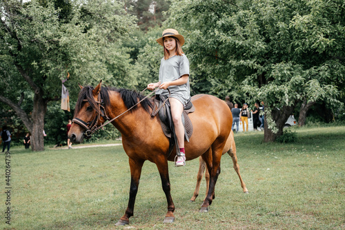 A happy young girl in a staw hat riding a brown horse in the countryside, a horse ranch in the summer © Marinesea
