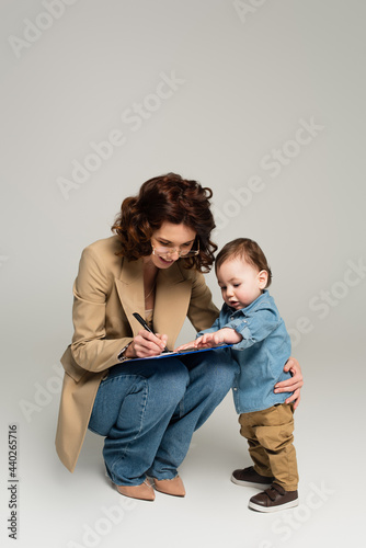 cheerful businesswoman in glasses writing on clipboard near toddler son on grey
