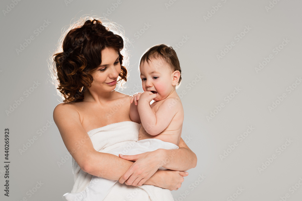 curly mother wrapped in towel holding baby boy in arms isolated on grey