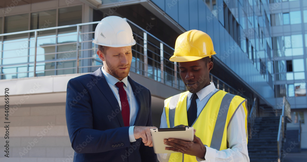 Diverse businessman and architect in hardhats with tablet pc computer at construction site Diverse businessman and architect in hardhats with tablet pc computer at construction site