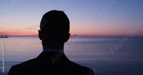 Businessman in formal black suit stands on beach and admires boundless sea with orange sunset reflections close view photo