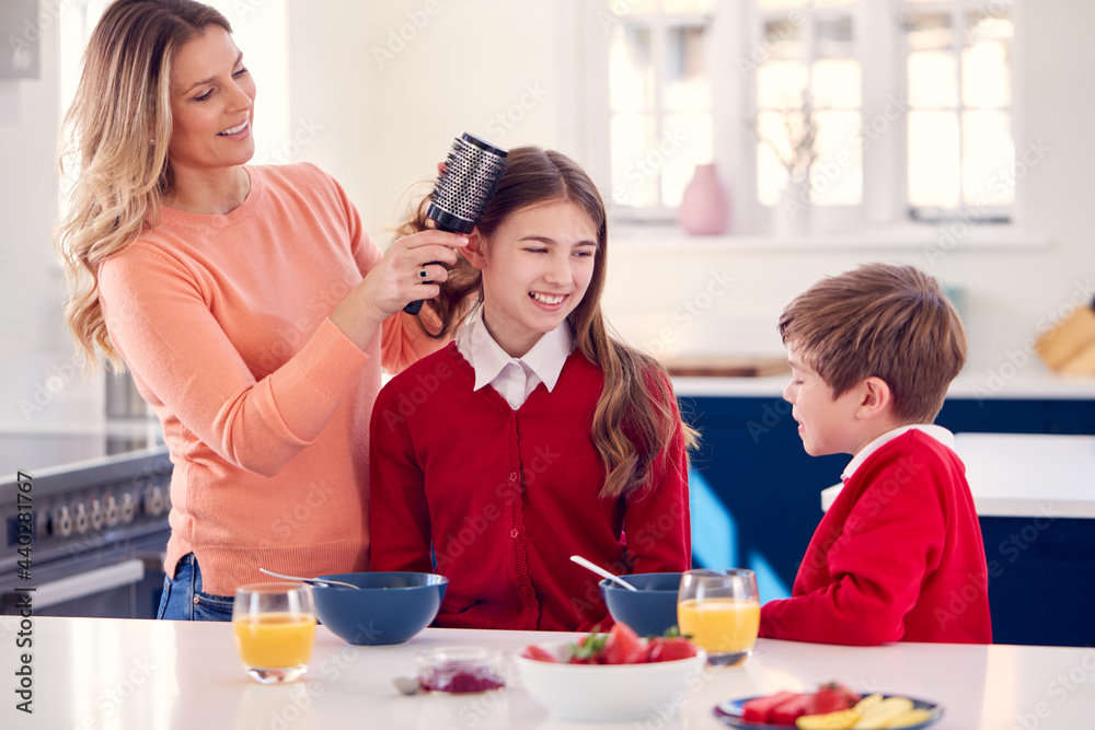 Mother Brushes Daughter's Hair As Children In School Uniform Eat Breakfast At Home Mother Brushes Daughter's Hair As Children In School Uniform Eat Breakfast At Home