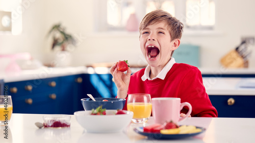 Laughing Boy Wearing School Uniform In Kitchen Eating Healthy Breakfast With Fresh Strawberry