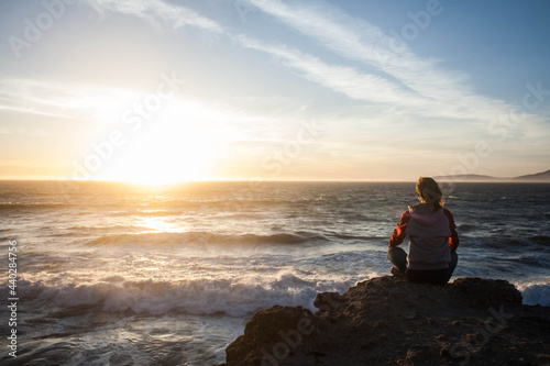 girl sitting in lotus position and meditating on a rock at sunset and looking at the ocean © Anastasiia