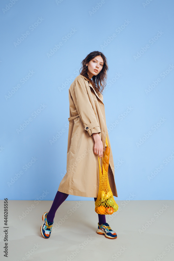 A young fashion girl in a beige trench, yellow short skirt, purple tights and sunglasses holds a mesh reusable eco bag with food. A young fashion girl in a beige trench, yellow short skirt, purple tights and sunglasses holds a mesh reusable eco bag with food.